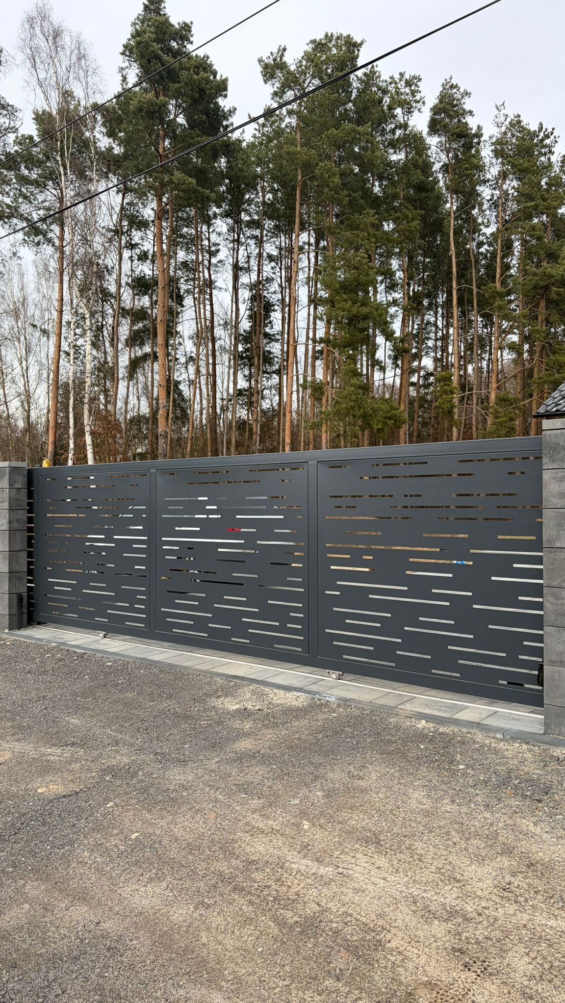 Nowoczesna, przesuwna brama w kolorze grafitowym z poziomymi, ażurowymi wycięciami, osadzona na betonowych słupkach. Tło stanowią wysokie drzewa.