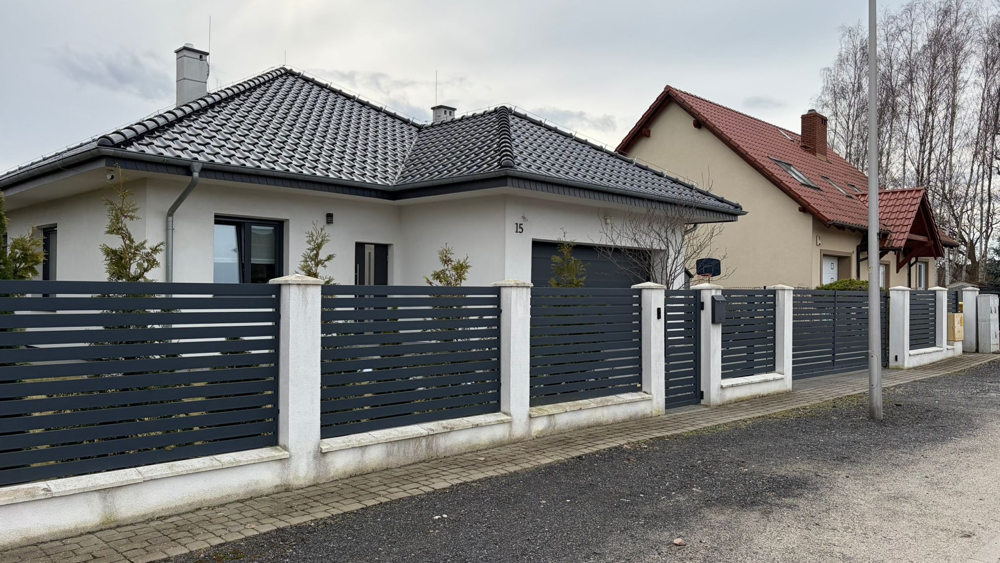 Nowoczesne ogrodzenie panelowe w kolorze grafitowym z białymi słupkami, otaczające posesję z domem jednorodzinnym. Widoczna brama wjazdowa i furtka.