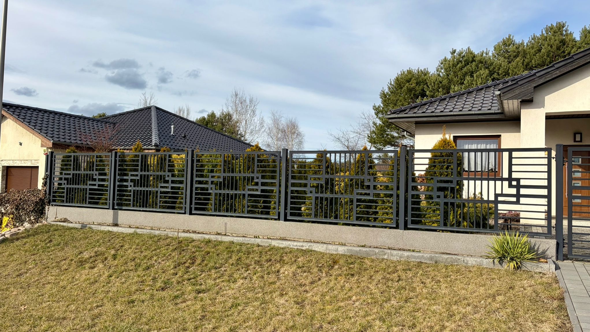 Nowoczesne, metalowe ogrodzenie posesji z geometrycznym wzorem, na tle domu z ciemną dachówką i zieleni. Widok z zewnątrz, w słoneczny dzień.