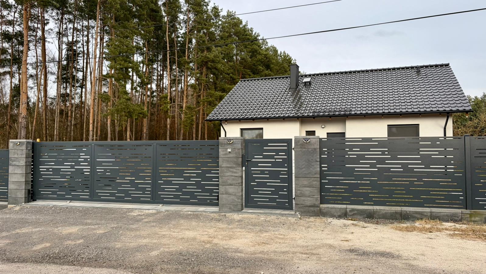 Nowoczesna, szara brama wjazdowa i przęsła ogrodzeniowe z poziomymi, ażurowymi wycięciami, osadzone na betonowych słupkach. W tle dom i las.