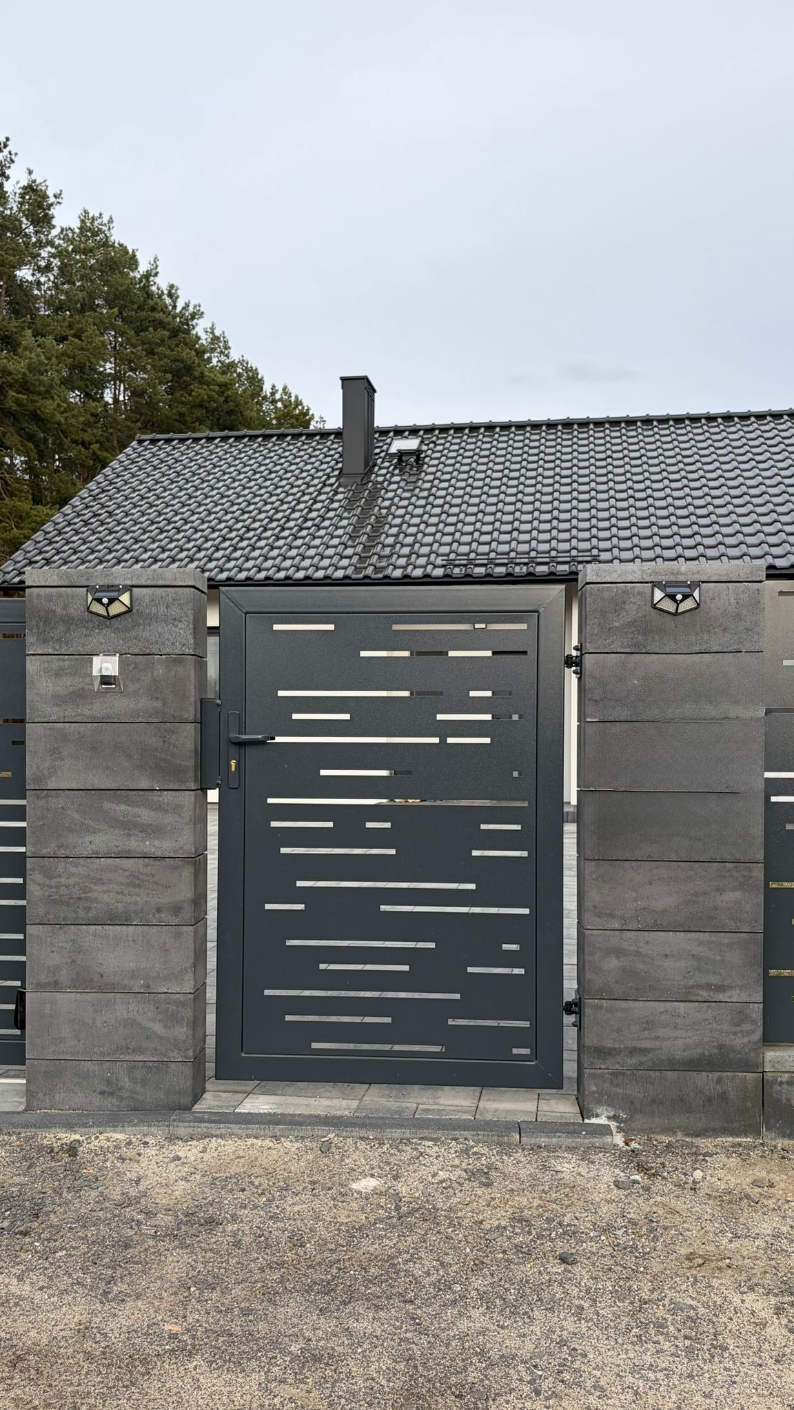 Nowoczesna, grafitowa brama wjazdowa z poziomymi, ażurowymi wycięciami, osadzona na betonowych słupkach z oświetleniem, na tle domu z ciemną dachówką.