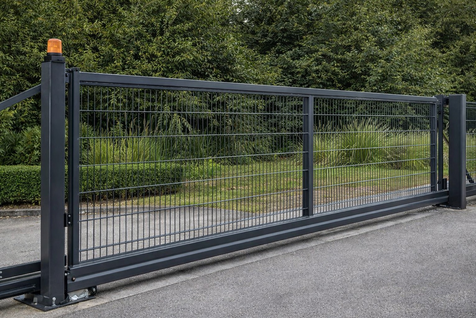 Przesuwna brama panelowa w kolorze grafitowym, z pomarańczową lampą ostrzegawczą na słupku. Widok z bliska na mechanizm jezdny i wypełnienie siatkowe na tle zieleni.