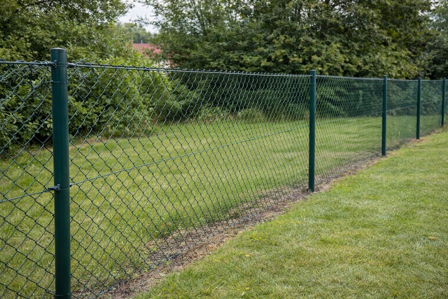 Zielona siatka ogrodzeniowa z metalowymi słupkami na trawniku, widok z boku. W tle drzewa i fragment dachu budynku.