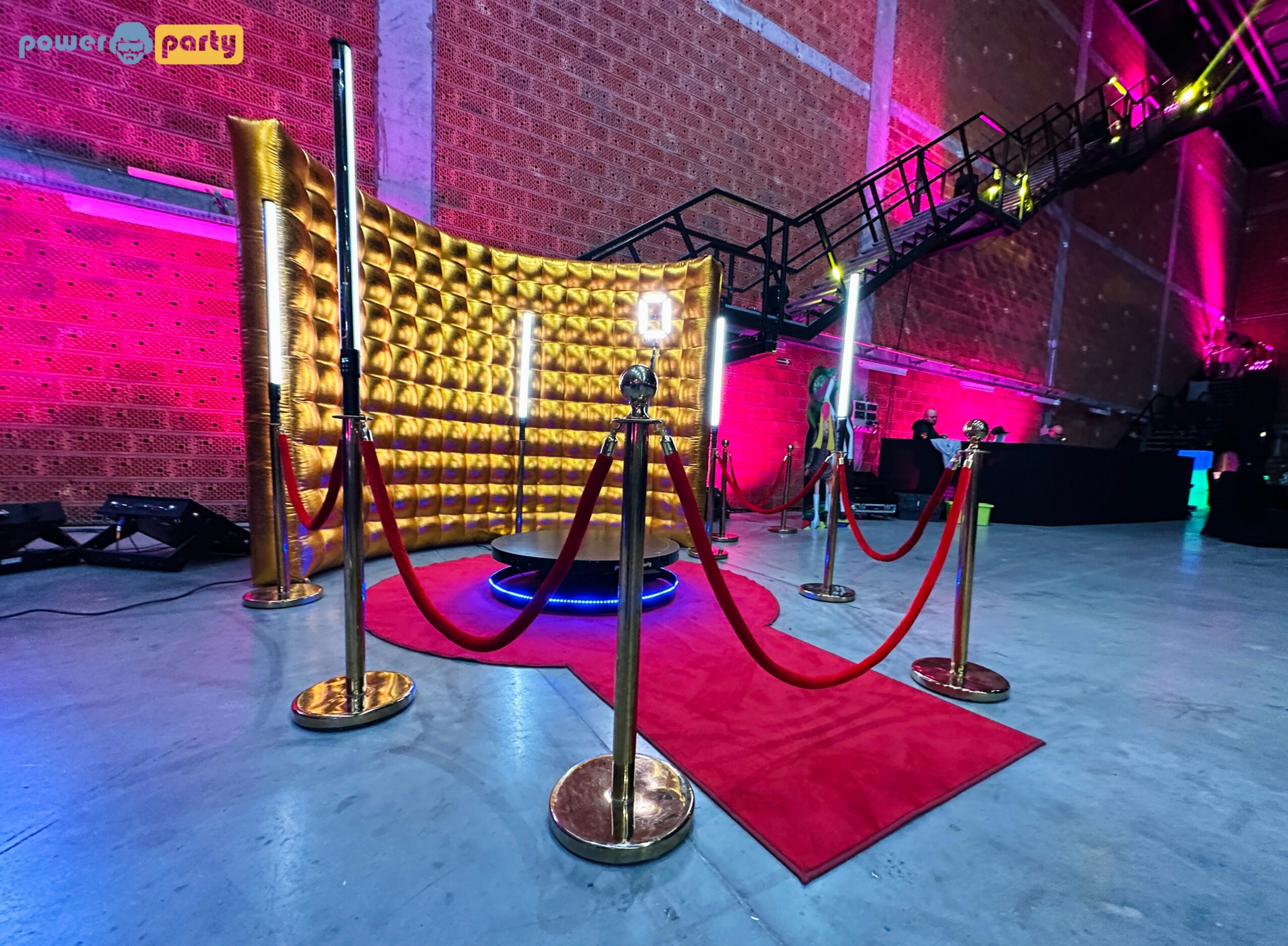 Złota ścianka z oświetleniem LED i czerwonym dywanem, otoczona słupkami z czerwonym sznurem, tworząca elegancką strefę do zdjęć na tle ceglanej ściany.