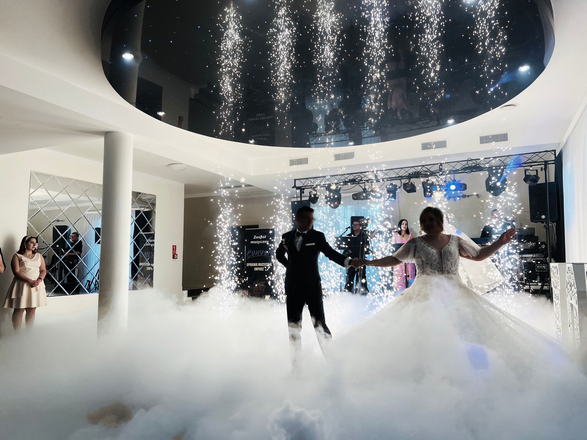 Para młoda w tańcu, otoczona dymem i fontannami iskier na sali weselnej z zespołem muzycznym w tle. Dekoracja sufitu z efektem lustra.
