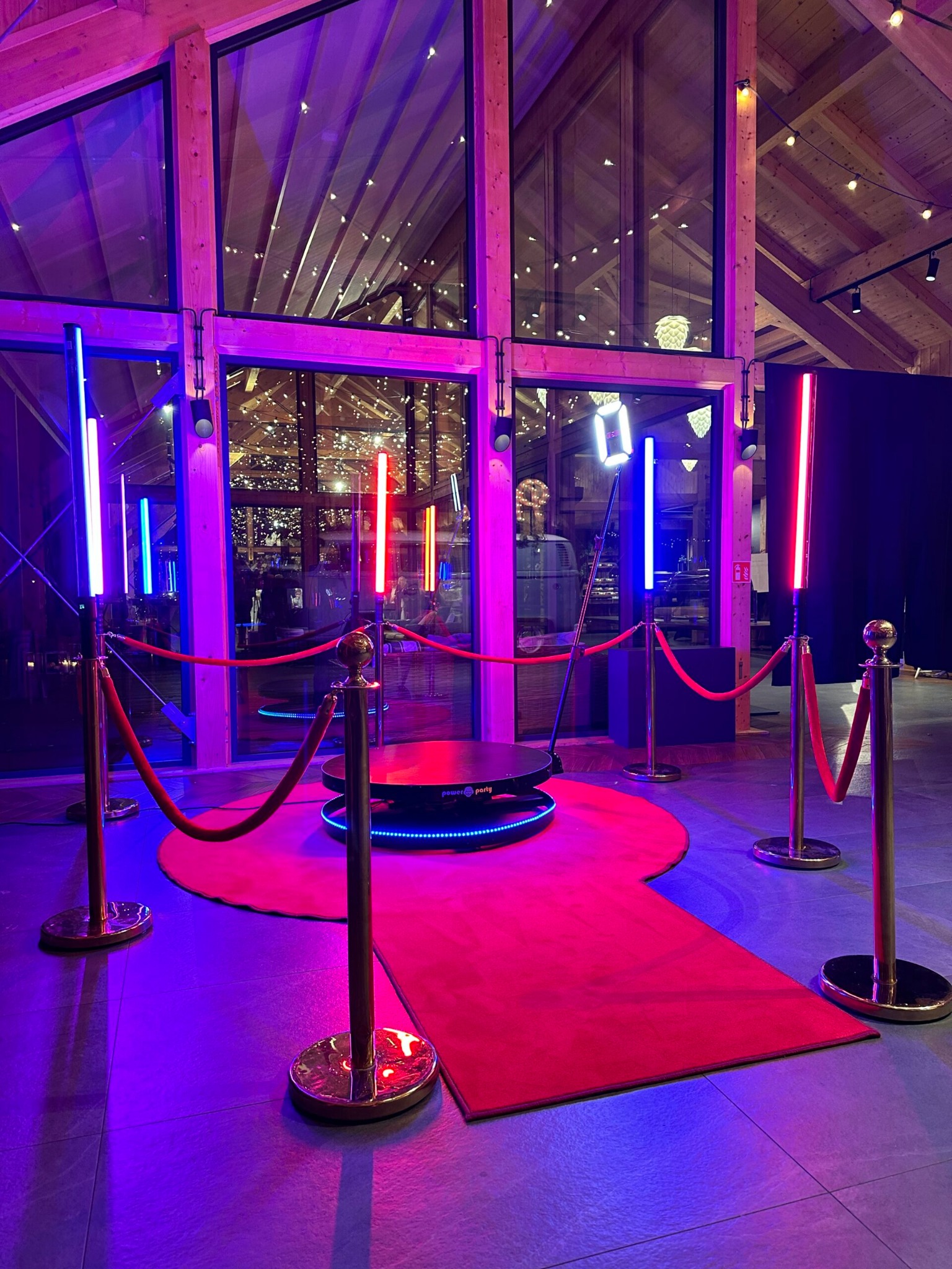 Rotacyjna fotobudka 360 z czerwonym dywanem i oświetleniem LED w kolorach niebieskim i czerwonym, ustawiona w przestronnym wnętrzu z drewnianą konstrukcją dachu.