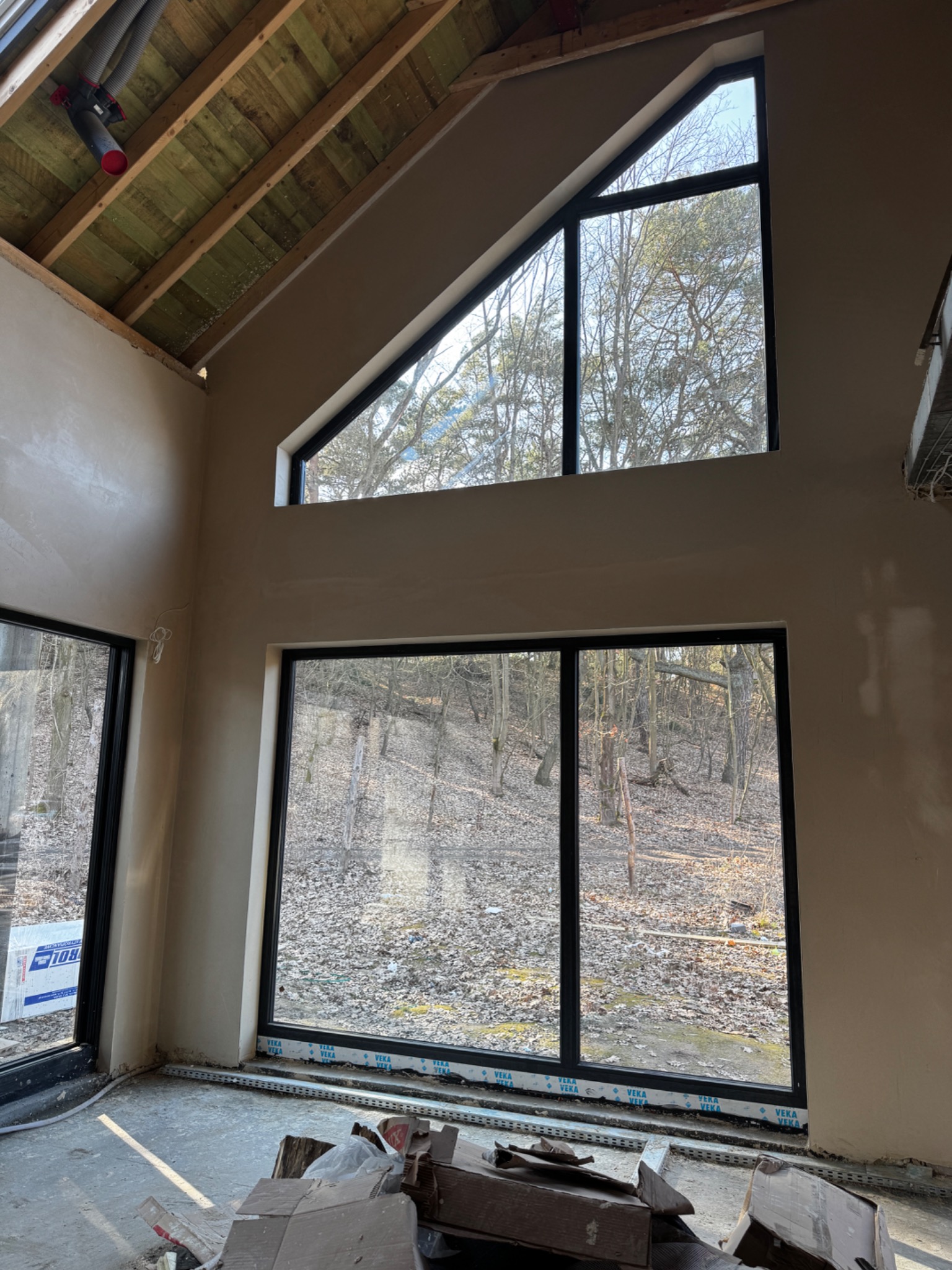 Wnętrze po tynkowaniu w Iwiecu: ściany pokryte świeżym tynkiem, duże okna z widokiem na las, surowy drewniany strop. Widoczne ślady prac wykończeniowych.
