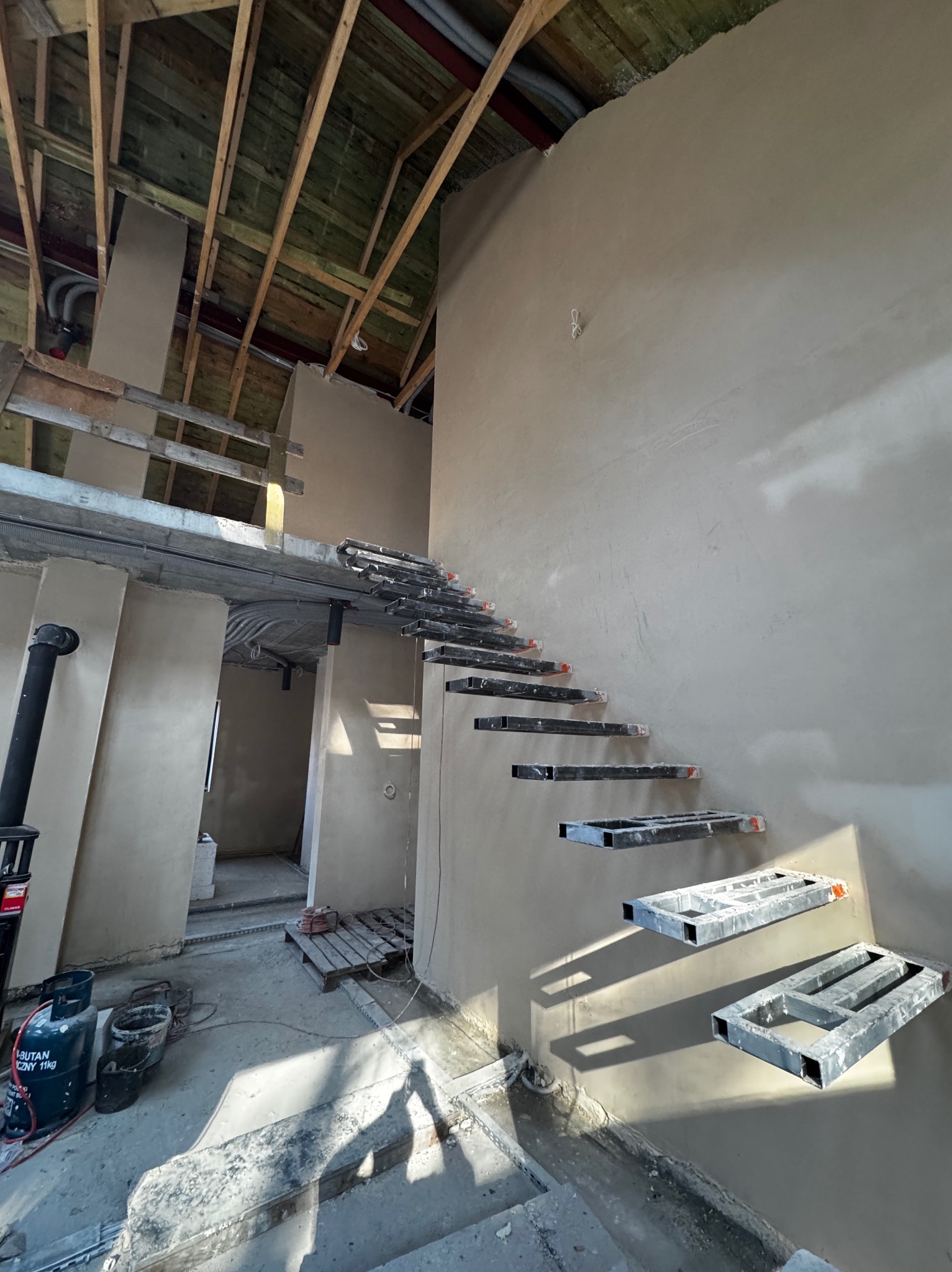 Wnętrze w trakcie prac wykończeniowych: otynkowane ściany, surowy strop drewniany i metalowe elementy schodów wiszących. Widoczne instalacje i narzędzia.