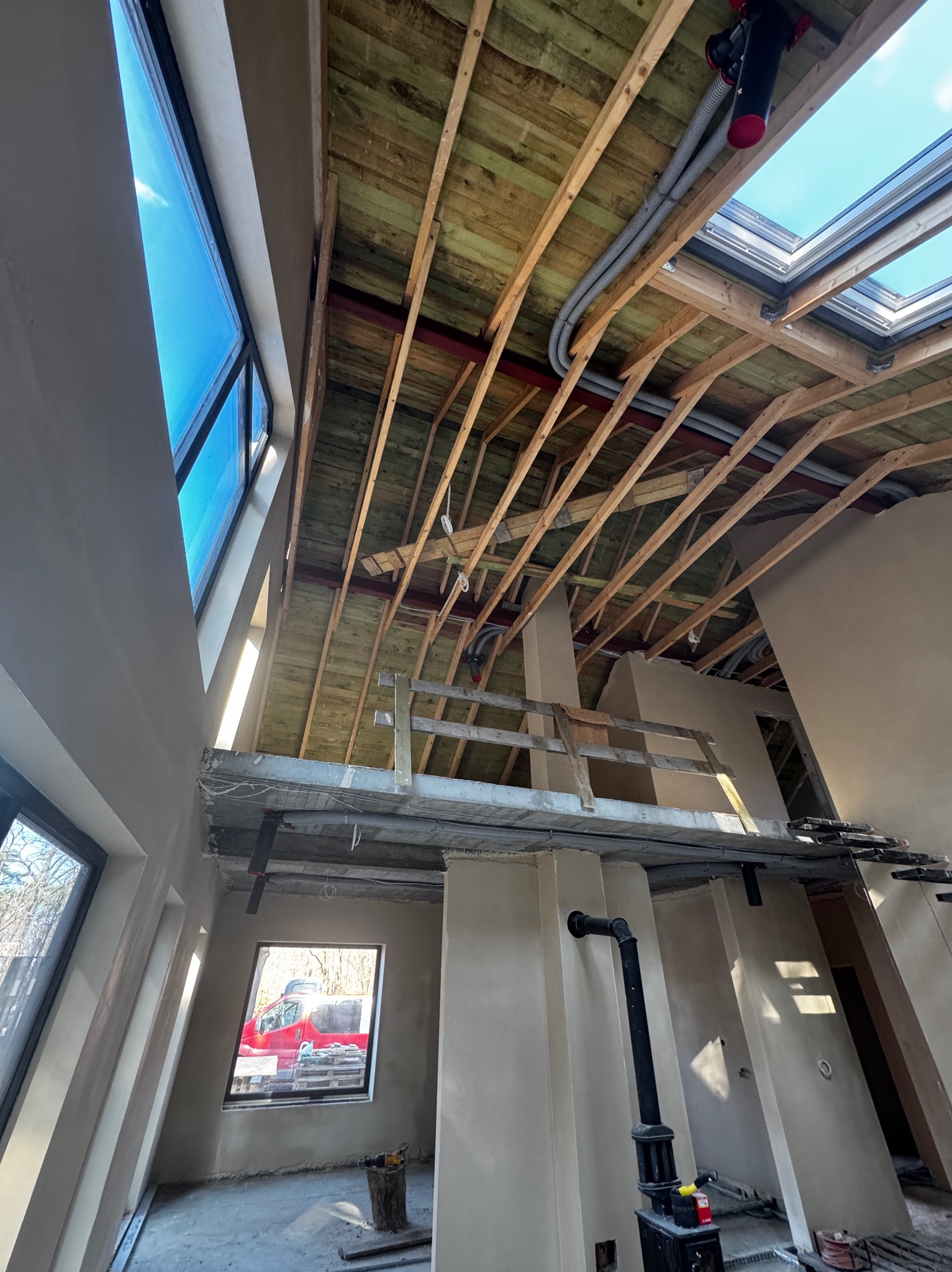 Wnętrze w trakcie prac wykończeniowych: surowy tynk na ścianach, drewniana konstrukcja dachu z oknami dachowymi, widoczne instalacje i rusztowanie. W oknach odbija się krajobraz.