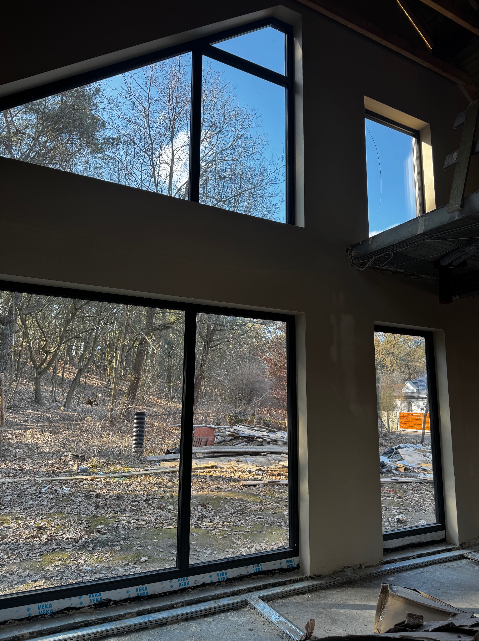 Surowe wnętrze z dużymi oknami z czarnymi ramami, widok na zewnątrz z drzewami bez liści. Ściany otynkowane, ale jeszcze nie wykończone. Elementy konstrukcyjne na podłodze.
