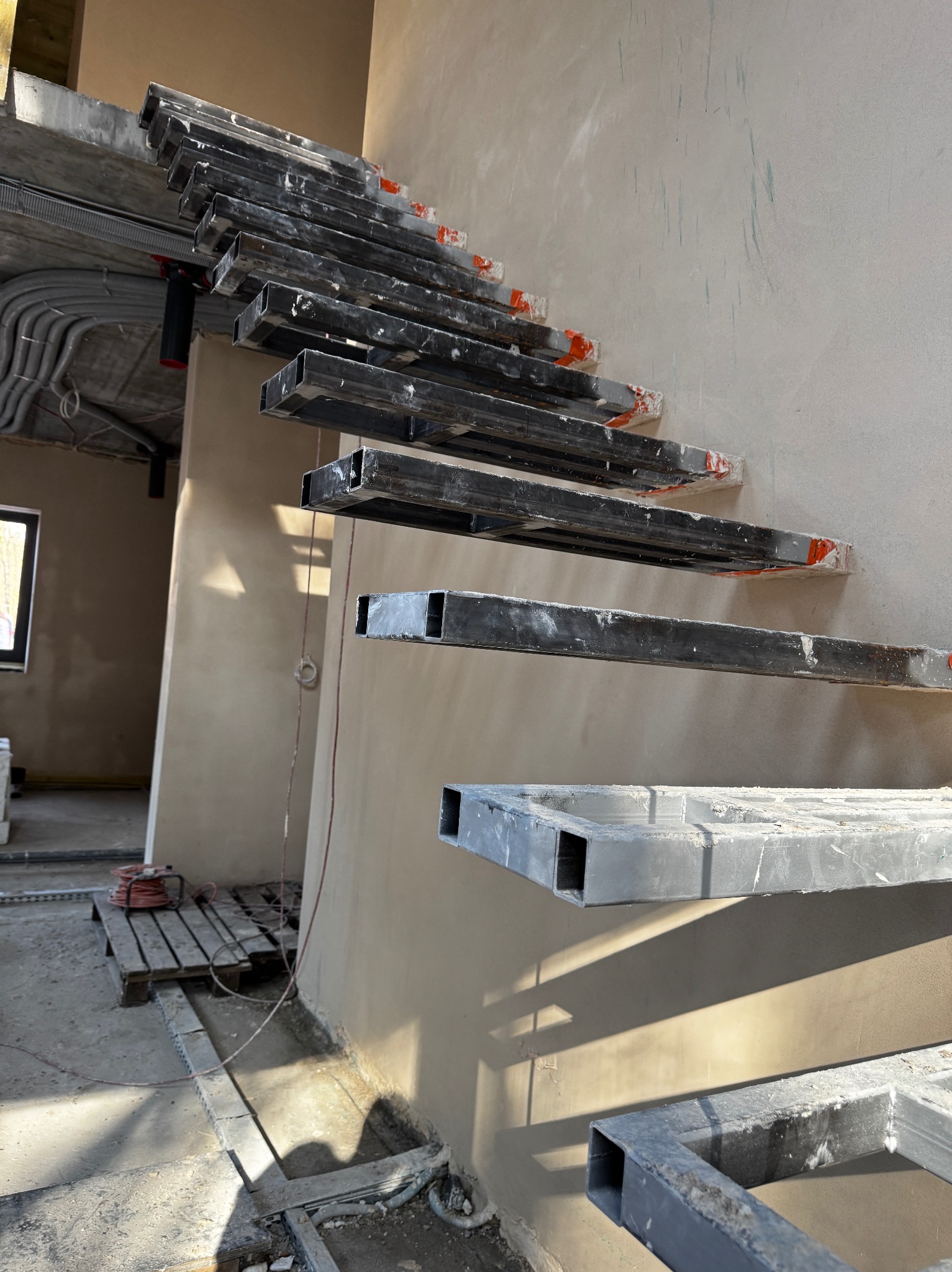 Surowe, metalowe stopnie schodów przymocowane do świeżo otynkowanej ściany w surowym wnętrzu. Widoczne instalacje elektryczne i narzędzia, co wskazuje na etap prac wykończeniowych.
