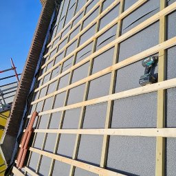 Dachy Elewacje Brodnica - Montaż drewnianej konstrukcji na ocieplonej szarej elewacji budynku, widoczny komin i narzędzia pracy. Ujęcie z dołu, błękitne niebo w tle.