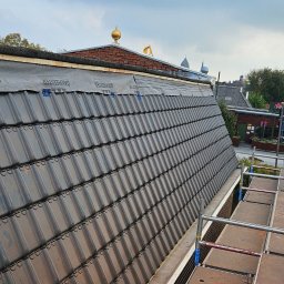 Dachy Elewacje Brodnica - Częściowy widok dachu z czarną dachówką, zabezpieczonego membraną, z rusztowaniem w tle i elementami zabudowy. Ujęcie z góry, w dzień.