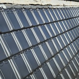 Dachy Elewacje Brodnica - Nowy, błyszczący dach z czarnej dachówki ceramicznej marki Koramic, ułożony pod kątem. Widoczne detale łączeń i fragment rusztowania. Tło rozmyte.