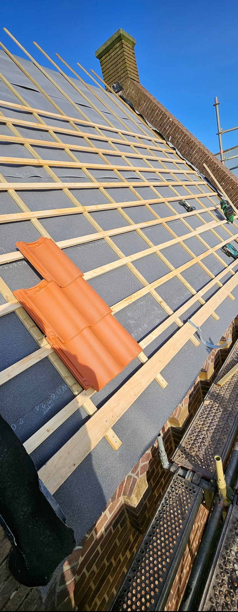 Montaż łat i kontrłat na membranie dachowej, przygotowanie pod dachówkę ceramiczną. Widoczny komin z cegły i fragment rusztowania.