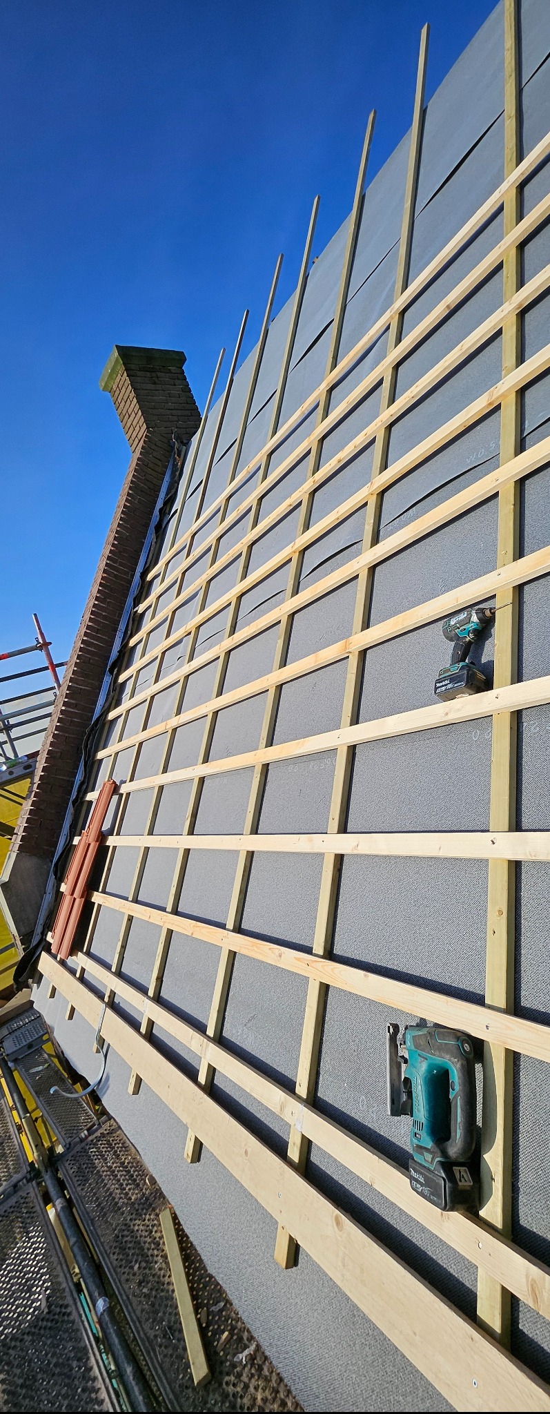 Montaż drewnianej konstrukcji na ocieplonej szarej elewacji budynku, widoczny komin i narzędzia pracy. Ujęcie z dołu, błękitne niebo w tle.