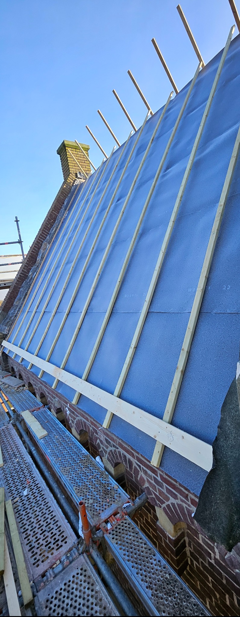 Skośny widok na dach w trakcie budowy: membrana dachowa, łaty, komin z cegły, rusztowanie. Błękitne niebo w tle. Widoczne detale konstrukcyjne.