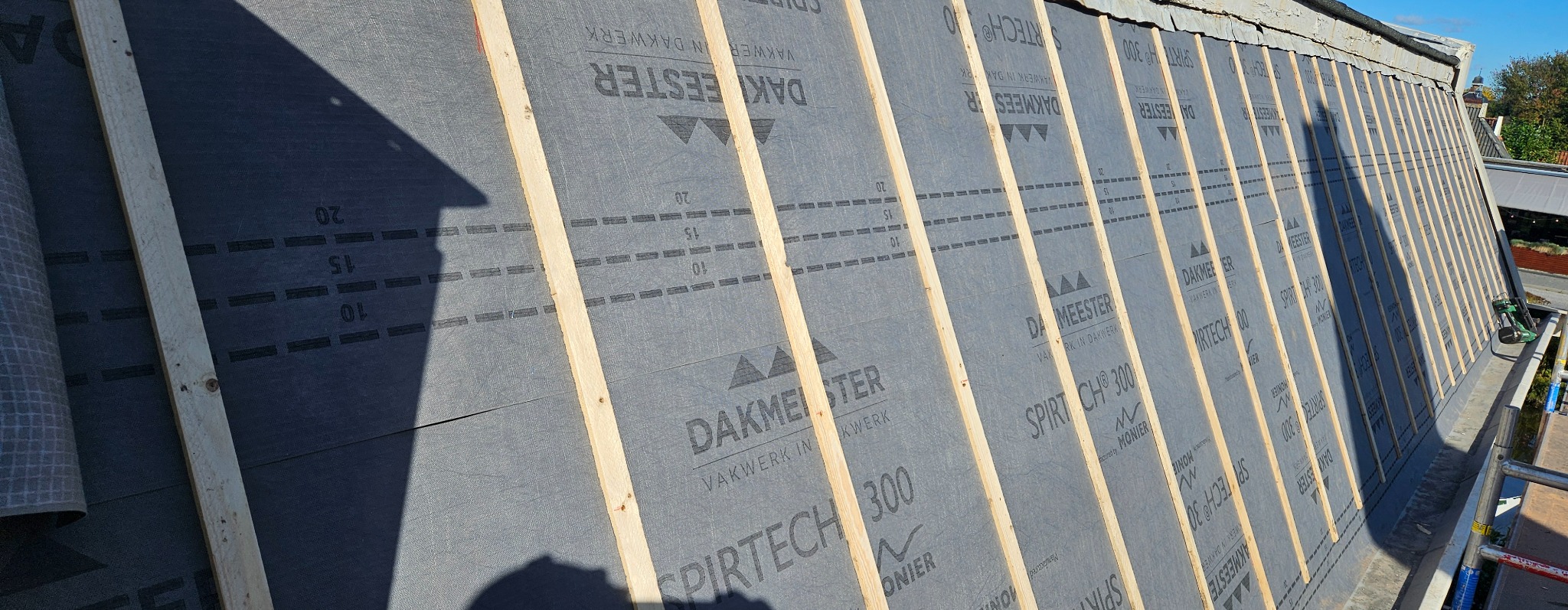 Dach w trakcie krycia: drewniane łaty na membranie dachowej z nadrukiem, widoczne narzędzia dekarskie i fragment rynny. Ujęcie z góry, w tle zieleń drzew.