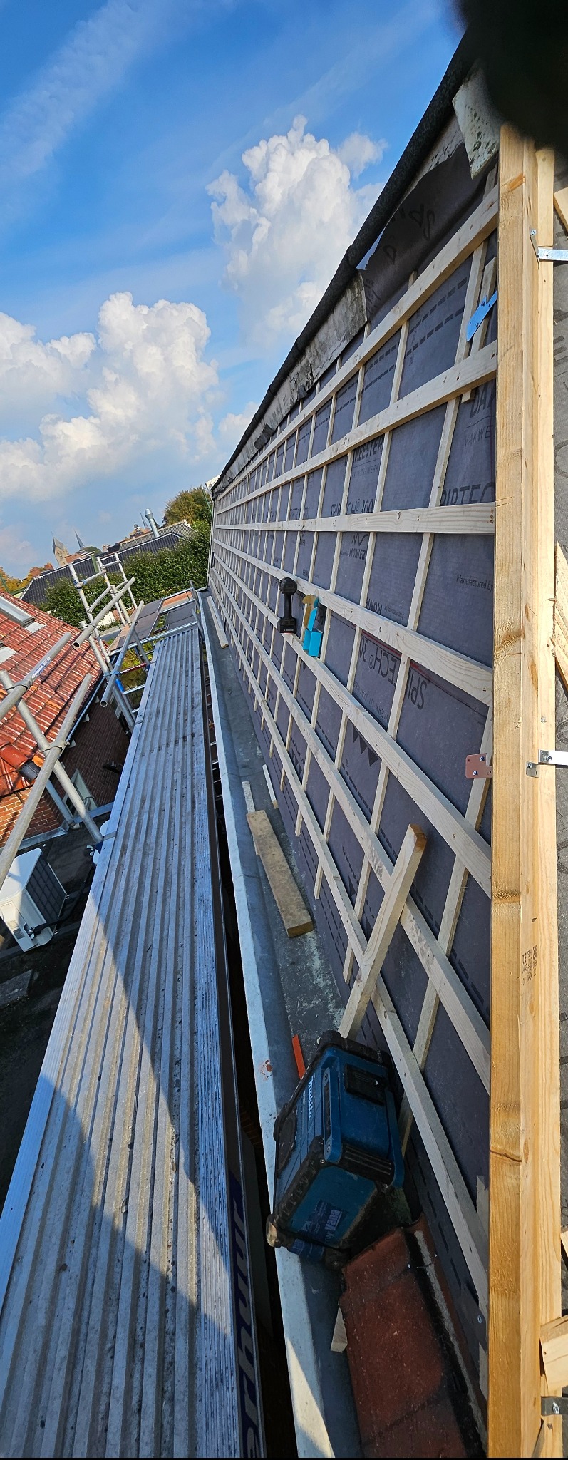 Montaż łat na dachu z membraną, widok z góry na rusztowanie i narzędzia. Jasne drewno kontrastuje z szarą membraną i błękitnym niebem.