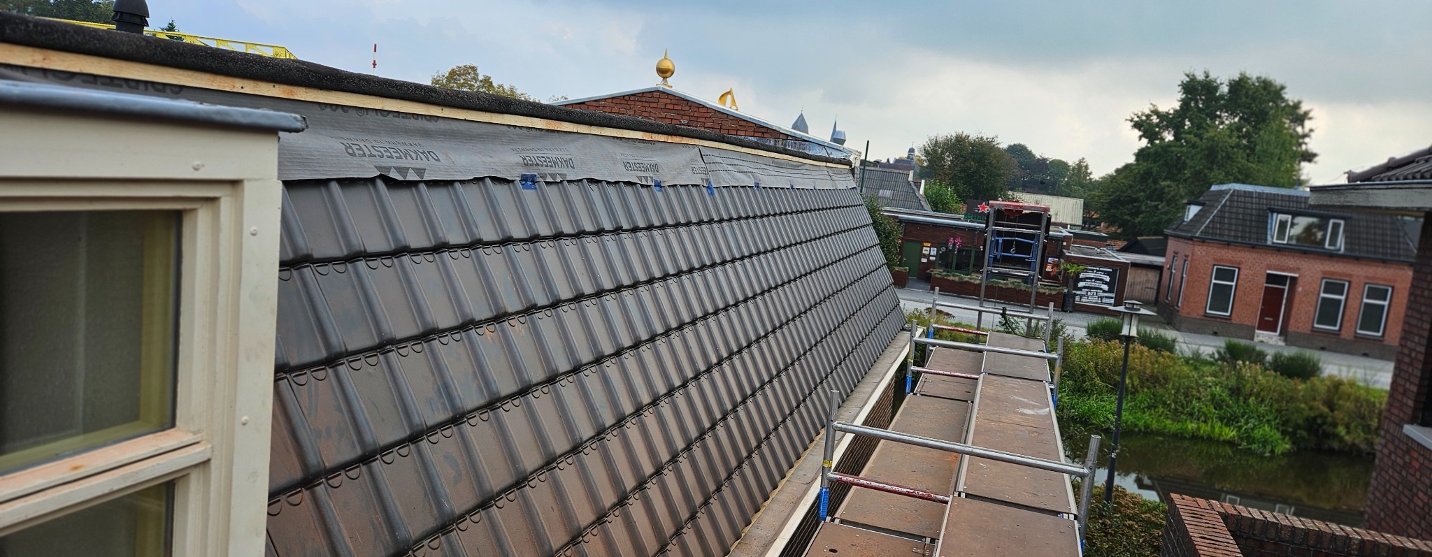 Częściowy widok dachu z czarną dachówką, zabezpieczonego membraną, z rusztowaniem w tle i elementami zabudowy. Ujęcie z góry, w dzień.