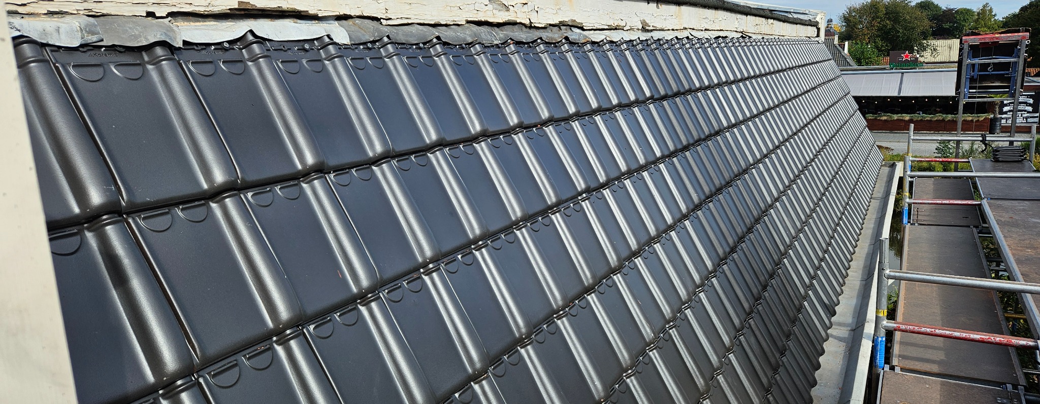 Nowy, błyszczący dach z czarnej dachówki ceramicznej marki Koramic, ułożony pod kątem. Widoczne detale łączeń i fragment rusztowania. Tło rozmyte.