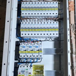 Kacper Brzezek - Rozdzielnica elektryczna w obudowie, z widocznymi bezpiecznikami, przewodami i złączkami. Elementy oznaczone etykietami. Widoczne fragmenty ściany z cegły.