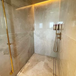 Ikona Lab Monika Stolarska - Nowoczesna kabina prysznicowa z beżowymi płytkami, złotą armaturą i subtelnym oświetleniem LED. Minimalistyczny design z odpływem liniowym.