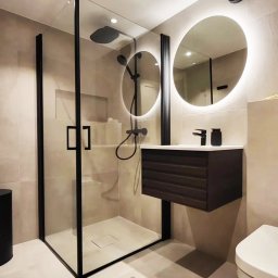 Ikona Lab Monika Stolarska - Nowoczesna łazienka z kabiną prysznicową typu walk-in z czarnymi profilami, okrągłym lustrem z podświetleniem LED i wiszącą szafką z umywalką w kolorze ciemnego drewna.