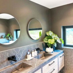 Ikona Lab Monika Stolarska - Nowoczesna łazienka z dwoma okrągłymi lustrami, marmurowym blatem z wpuszczonymi umywalkami i rośliną doniczkową. Minimalistyczny design, szare ściany i drewniane szafki.