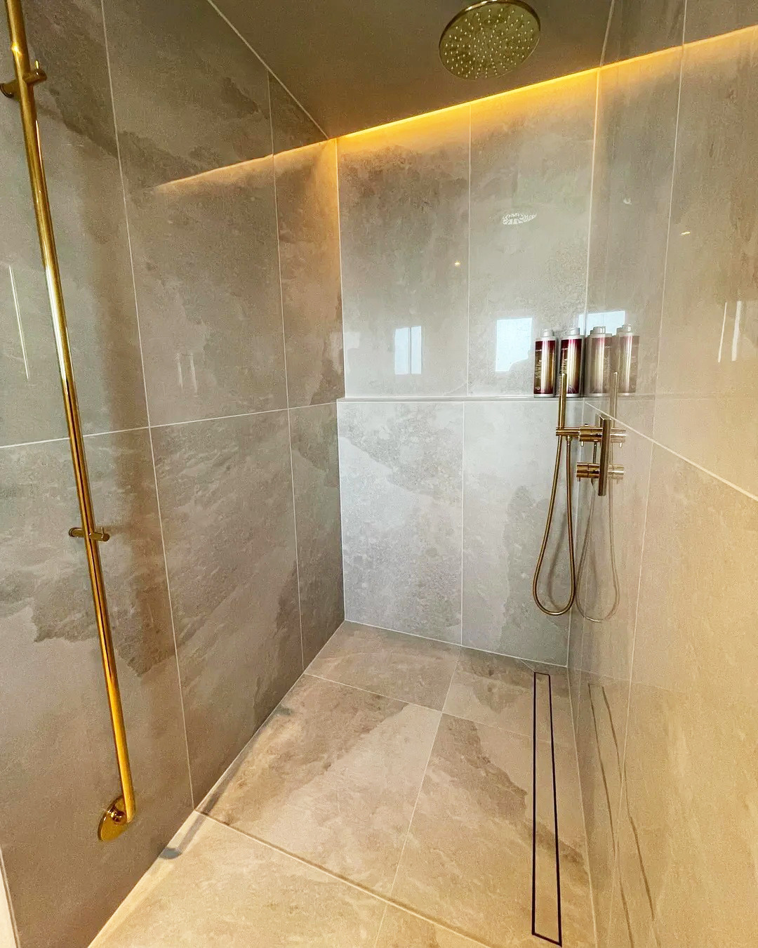 Nowoczesna kabina prysznicowa z beżowymi płytkami, złotą armaturą i subtelnym oświetleniem LED. Minimalistyczny design z odpływem liniowym.