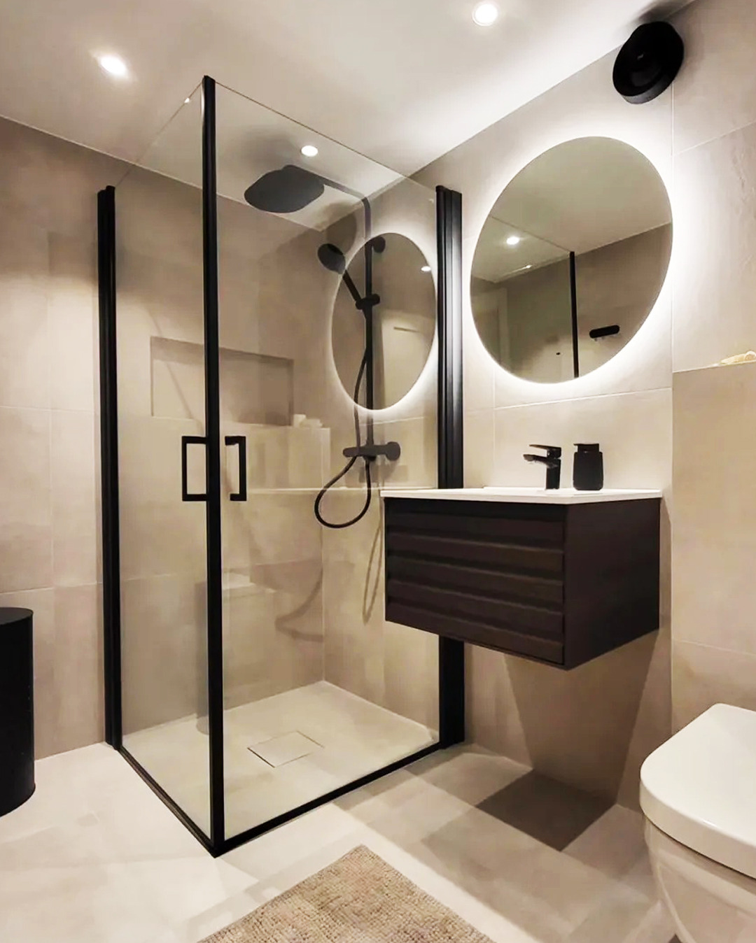 Nowoczesna łazienka z kabiną prysznicową typu walk-in z czarnymi profilami, okrągłym lustrem z podświetleniem LED i wiszącą szafką z umywalką w kolorze ciemnego drewna.