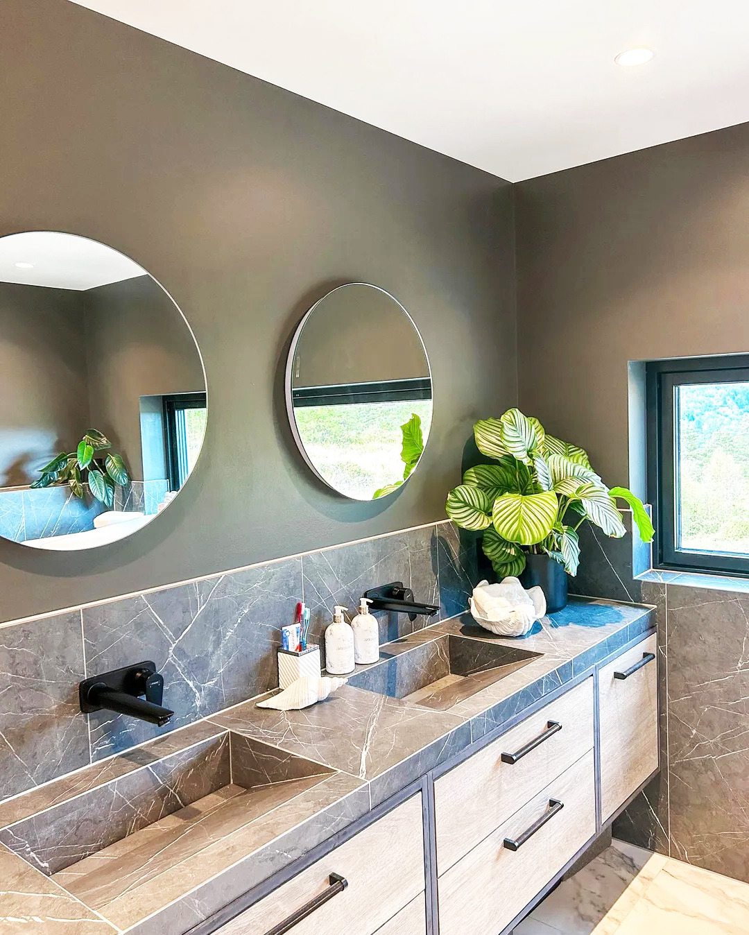 Nowoczesna łazienka z dwoma okrągłymi lustrami, marmurowym blatem z wpuszczonymi umywalkami i rośliną doniczkową. Minimalistyczny design, szare ściany i drewniane szafki.