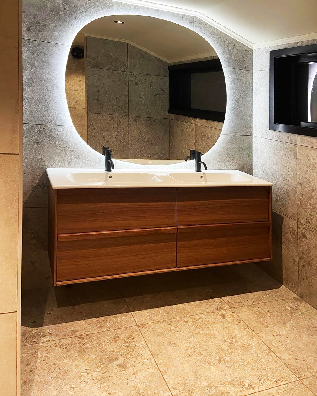 Nowoczesna łazienka: podwójna umywalka z drewnianą szafką, owalne lustro z podświetleniem LED, szare płytki na ścianach i beżowe na podłodze.