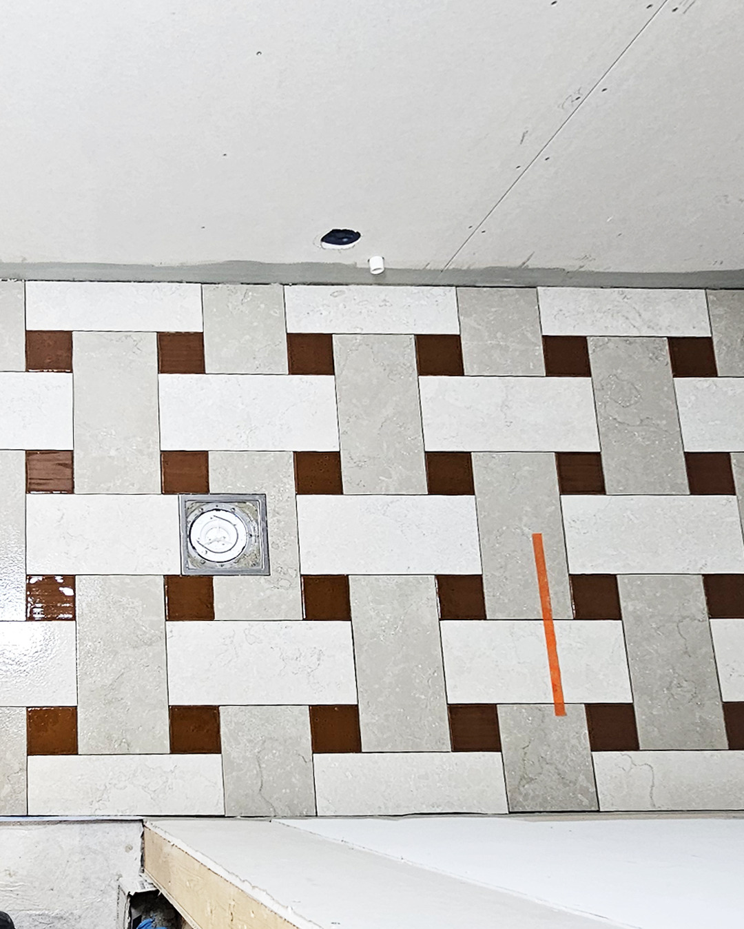 Ułożone płytki podłogowe w łazience, wzór w kratkę z brązowymi akcentami. Widoczny odpływ liniowy i fragment sufitu z otworem na oświetlenie.