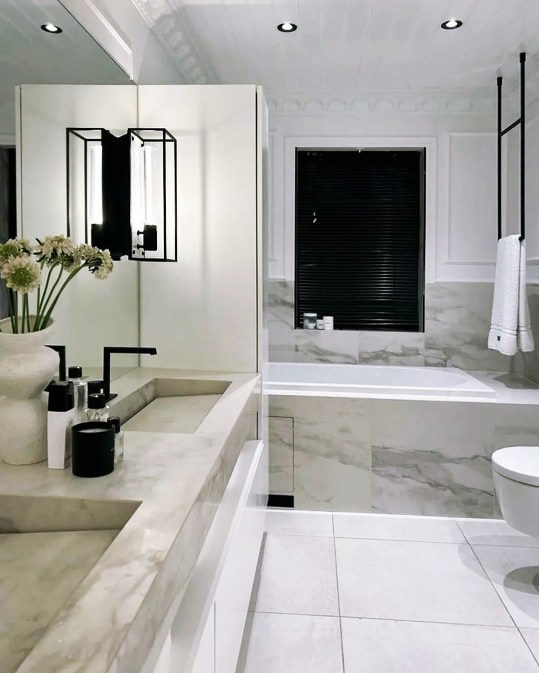 Elegancka łazienka w bieli i szarości z marmurowym blatem, czarną armaturą i geometrycznym oświetleniem. Minimalistyczny design z wanną i toaletą.