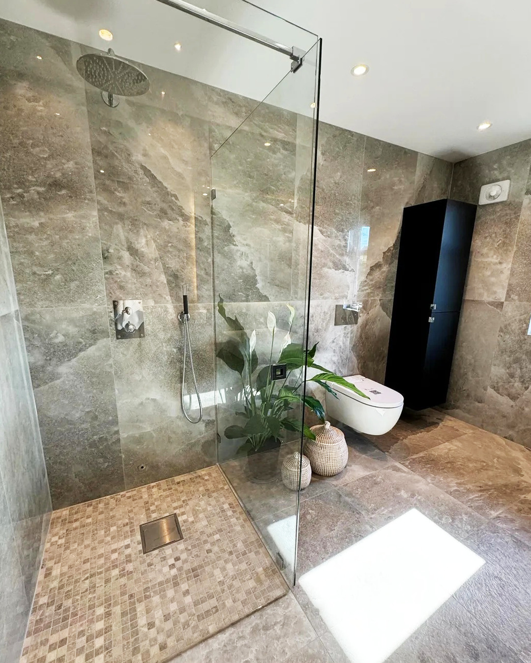 Nowoczesna łazienka z kabiną prysznicową typu walk-in, wykończona płytkami imitującymi marmur. Uwagę zwraca minimalistyczna toaleta i dekoracyjna roślina.