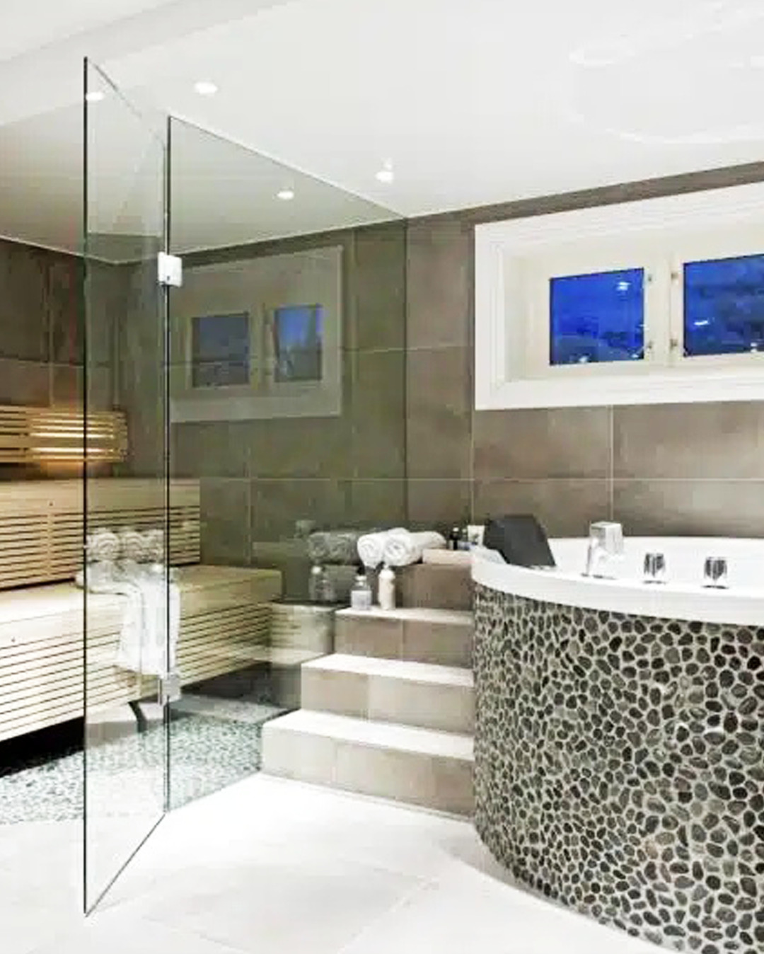 Nowoczesna łazienka z sauną, szklaną kabiną prysznicową i wanną z obudową z kamieni. Minimalistyczny design, stonowana kolorystyka, eleganckie wykończenie.