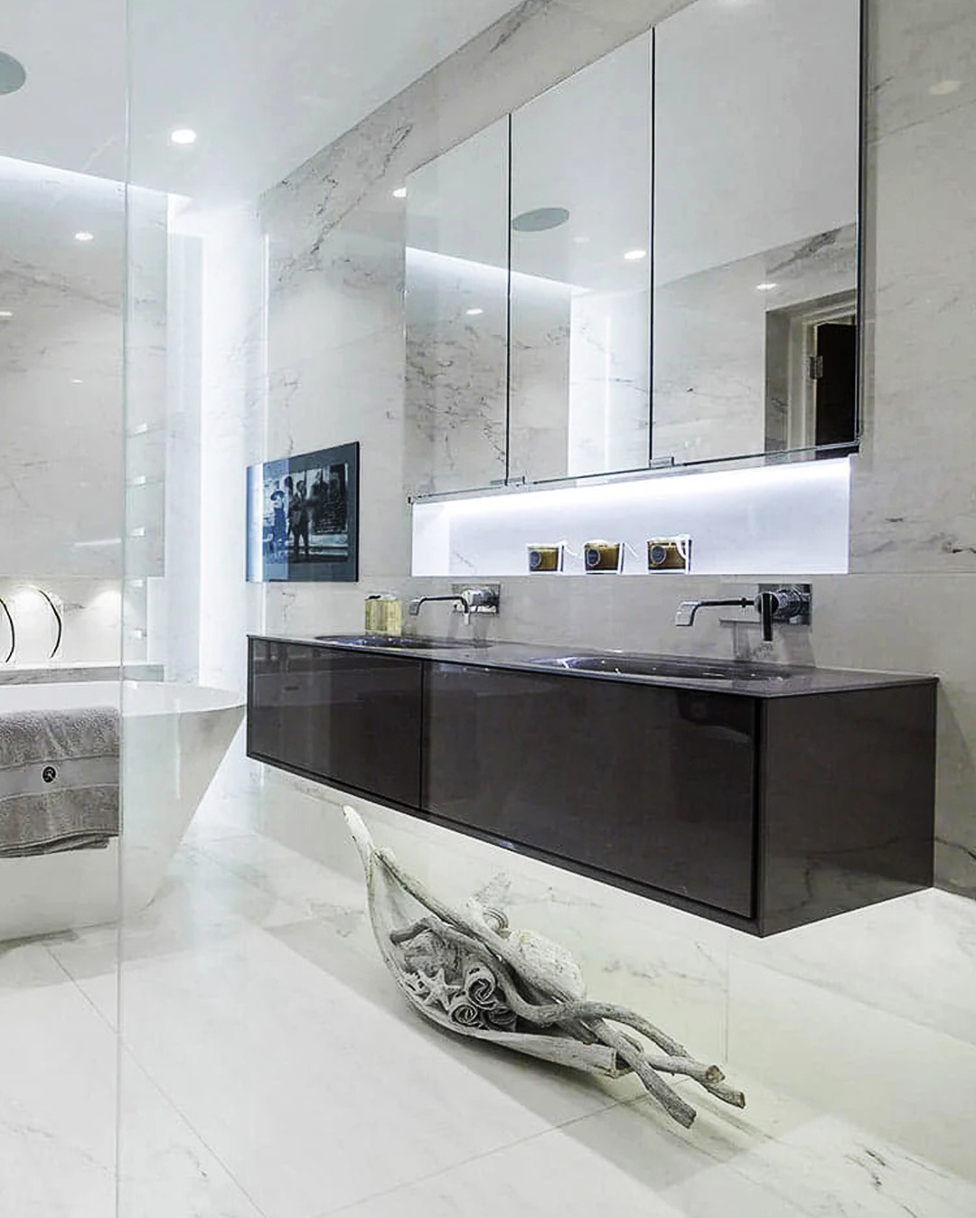 Elegancka łazienka z marmurowymi ścianami i podłogą, podwieszana szafka z umywalkami, duże lustro z oświetleniem LED i dekoracyjny element z drewna na podłodze.