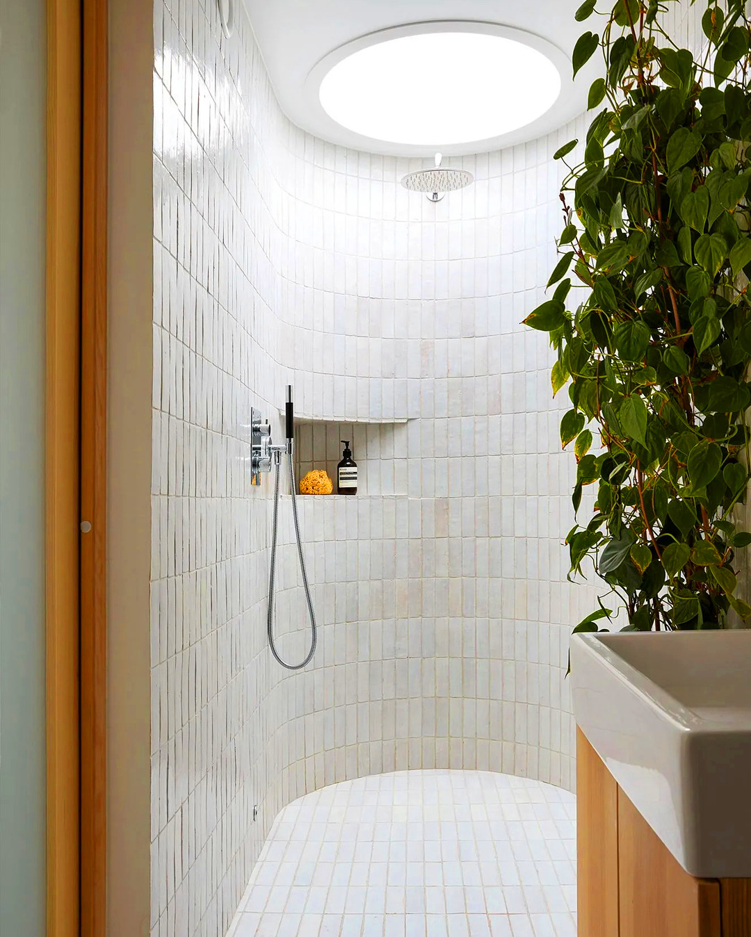Nowoczesna kabina prysznicowa z zaokrąglonymi ścianami wyłożonymi białymi, podłużnymi płytkami. Roślina doniczkowa dodaje naturalnego akcentu. Minimalistyczny design.
