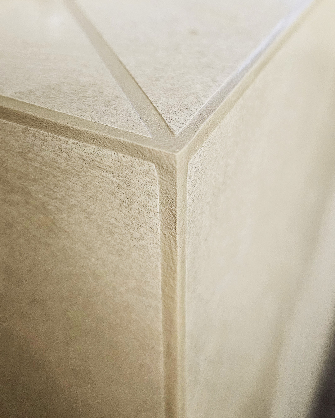Detal narożnika ściany z dekoracyjną, geometryczną fakturą. Jasny, neutralny odcień. Precyzyjne wykonanie, widoczne linie i krawędzie. Minimalistyczny design.
