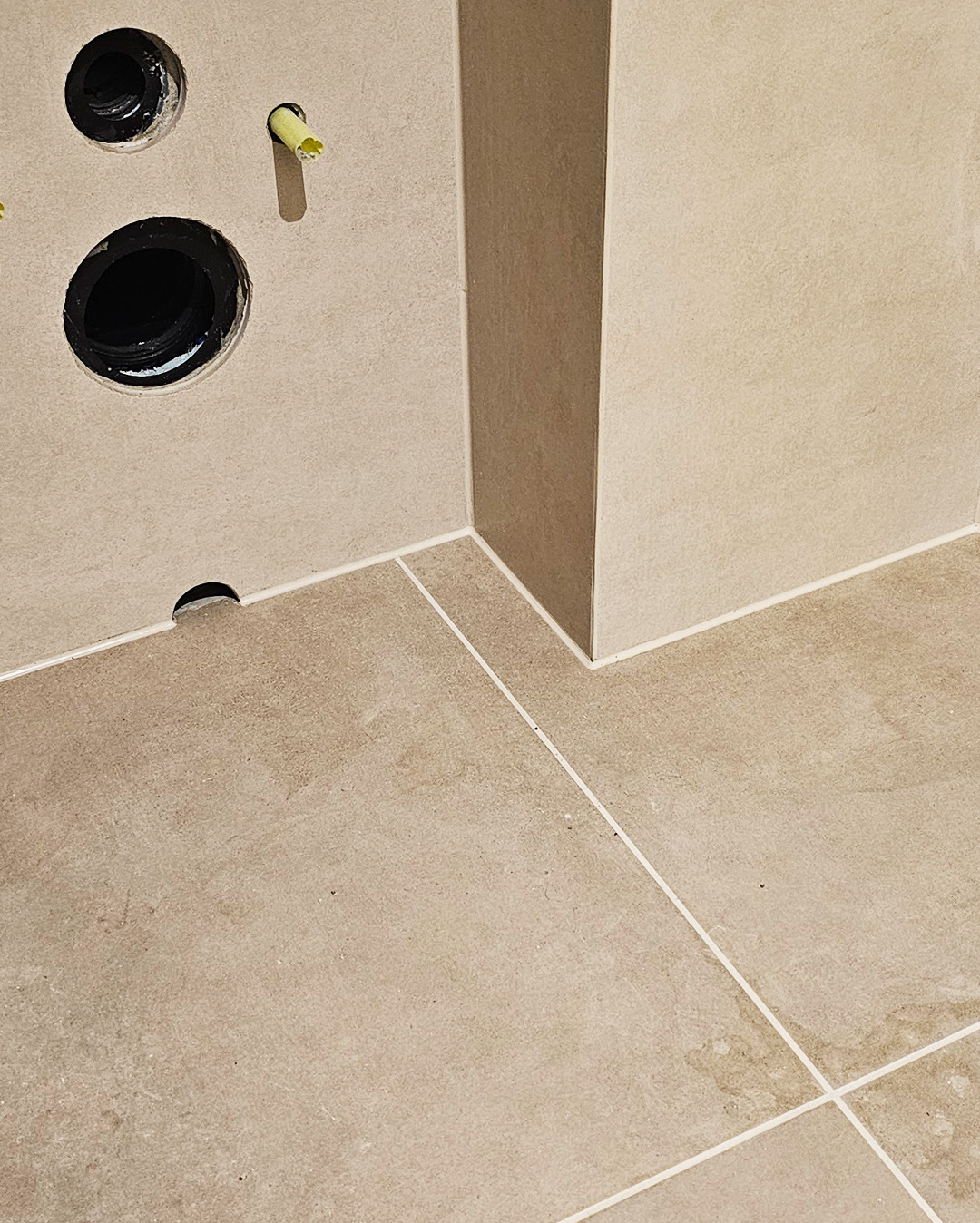 Beżowe płytki podłogowe i ścienne w łazience, widoczne otwory na instalacje. Minimalistyczne wykończenie z fugami w jasnym odcieniu. Ujęcie z bliska na łączenie płytek.