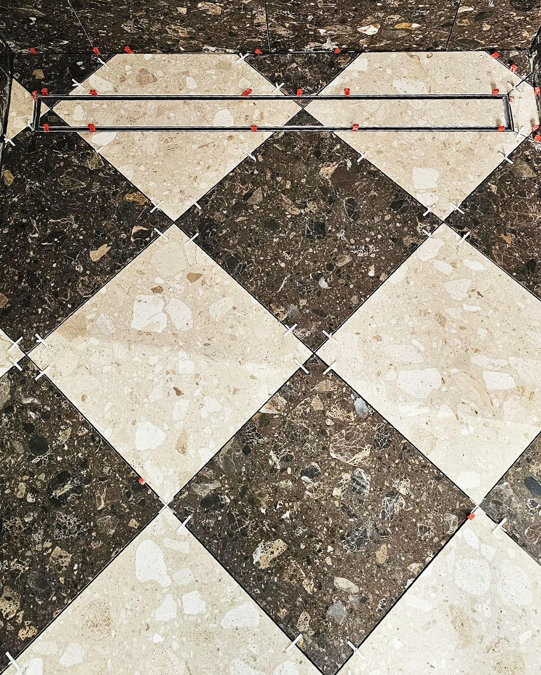 Ułożona podłoga w łazience z płytek w romby, beżowych i brązowych, z odpływem liniowym. Widoczne krzyżyki dystansowe.