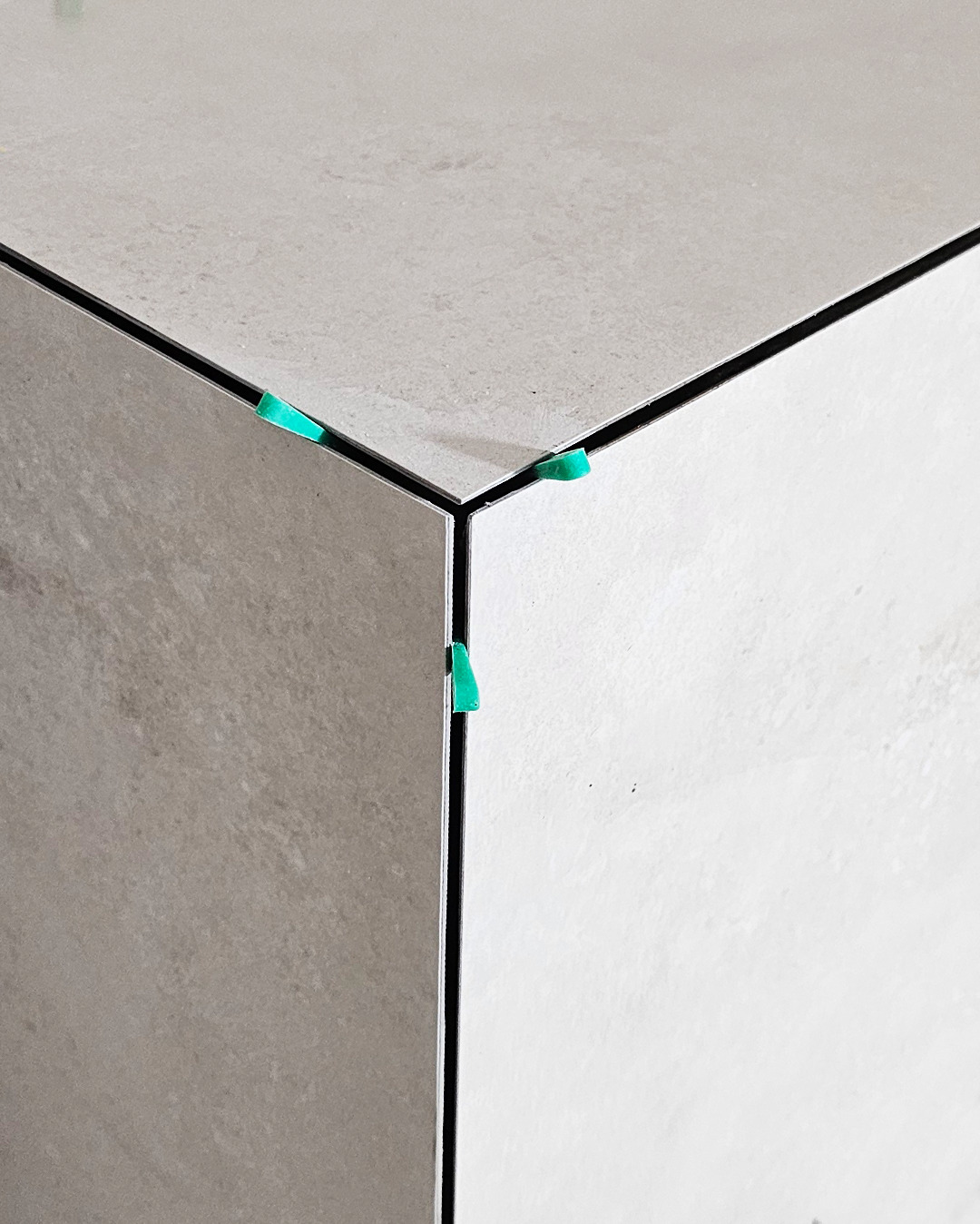 Ułożone płytki w rogu pomieszczenia, z użyciem zielonych klinów dystansowych, tworzące równe spoiny. Minimalistyczne ujęcie detalu wykończenia.