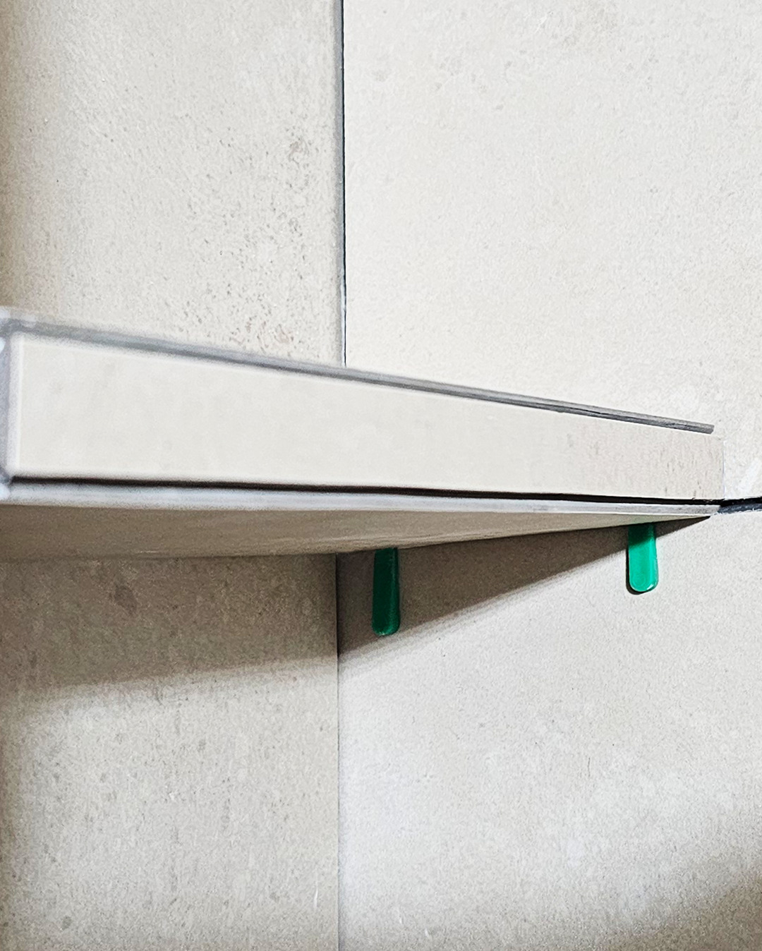 Fragment wykończonej ściany z jasnych płytek, z widocznymi zielonymi elementami poziomującymi. Ujęcie z bliska, ukazujące detale montażu płytek w rogu.