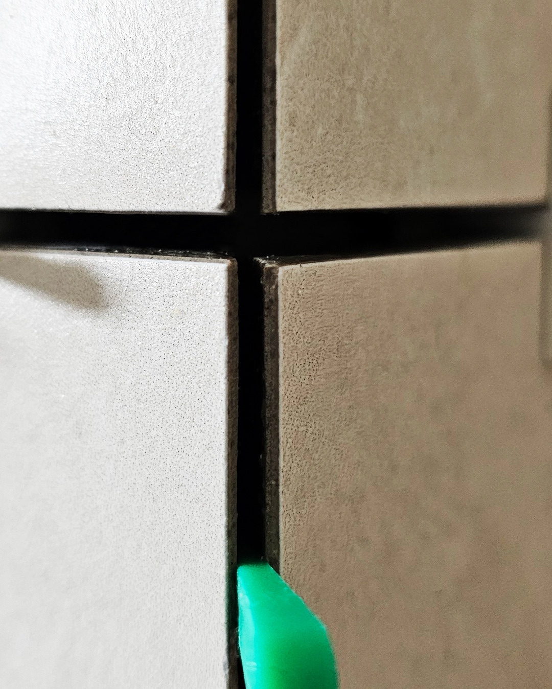 Beżowe płytki ceramiczne ułożone krzyżowo z ciemną fugą, z bliska widoczny fragment zielonego narzędzia do rozprowadzania fugi.