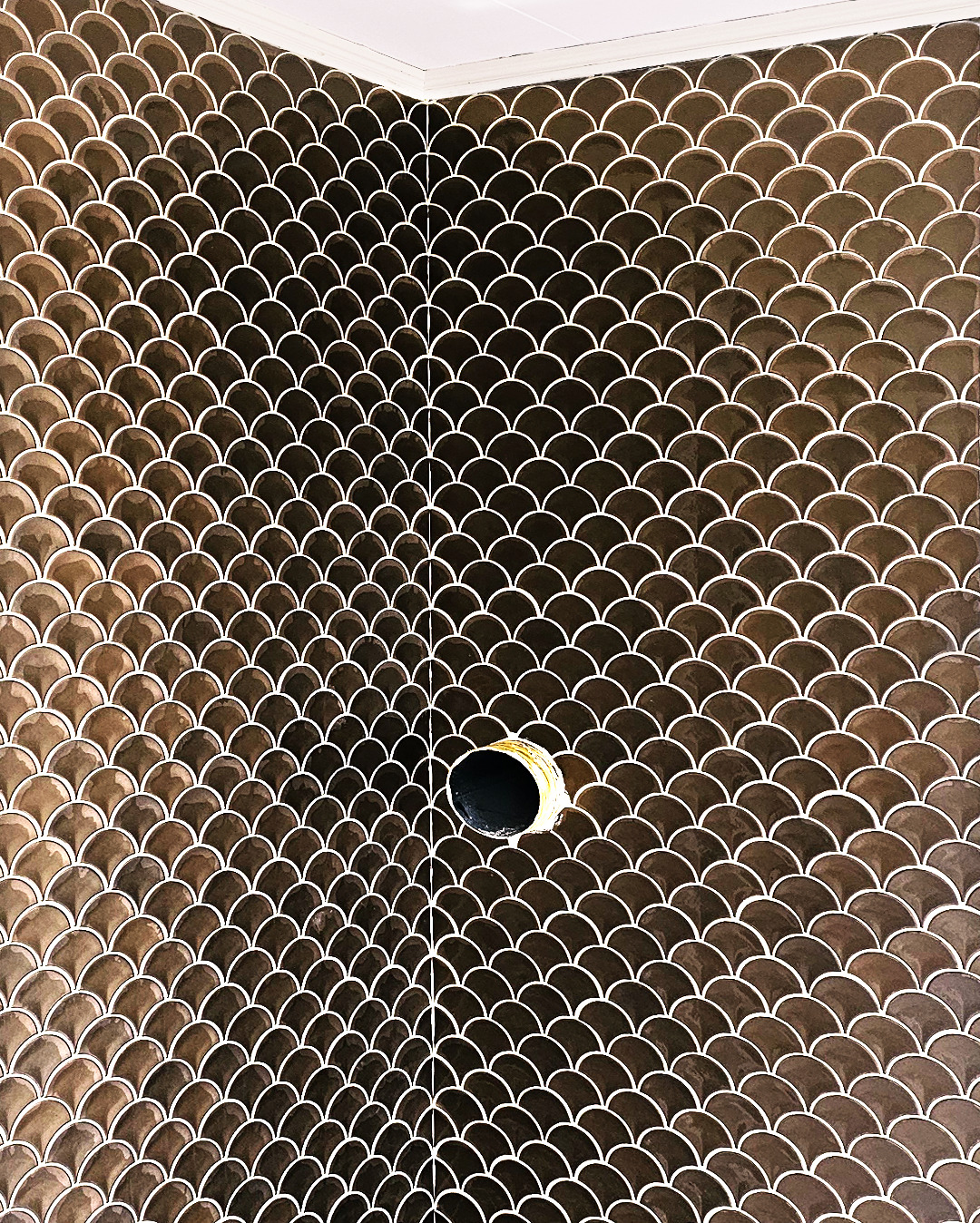 Brązowe płytki w kształcie rybiej łuski na ścianie łazienki z otworem na rurę. Nowoczesne wykończenie wnętrza, precyzyjne ułożenie glazury.