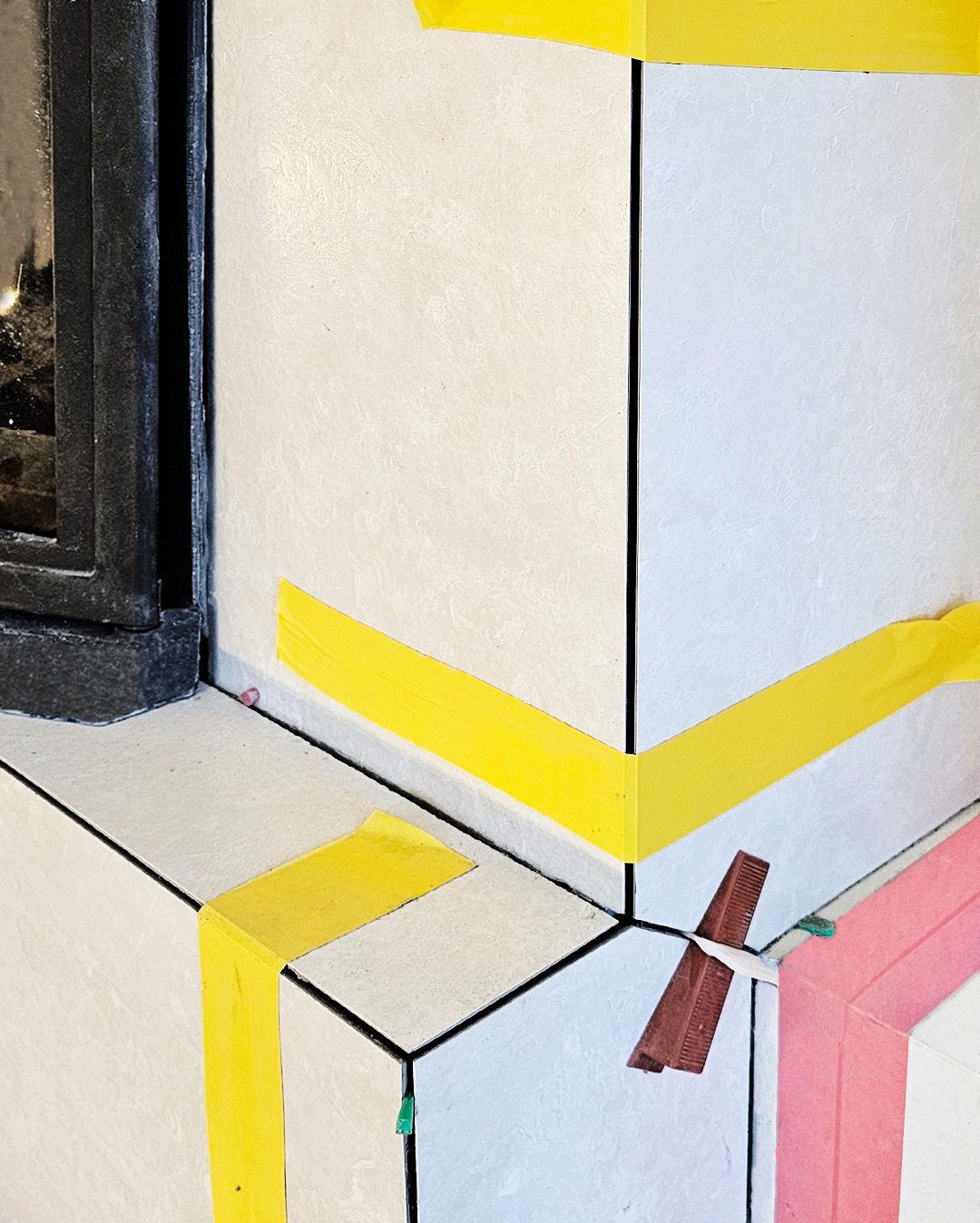 Układanie jasnych płytek przy obudowie kominka, zabezpieczonych żółtą taśmą i klinami. Widoczne fugi i detale wykończenia.