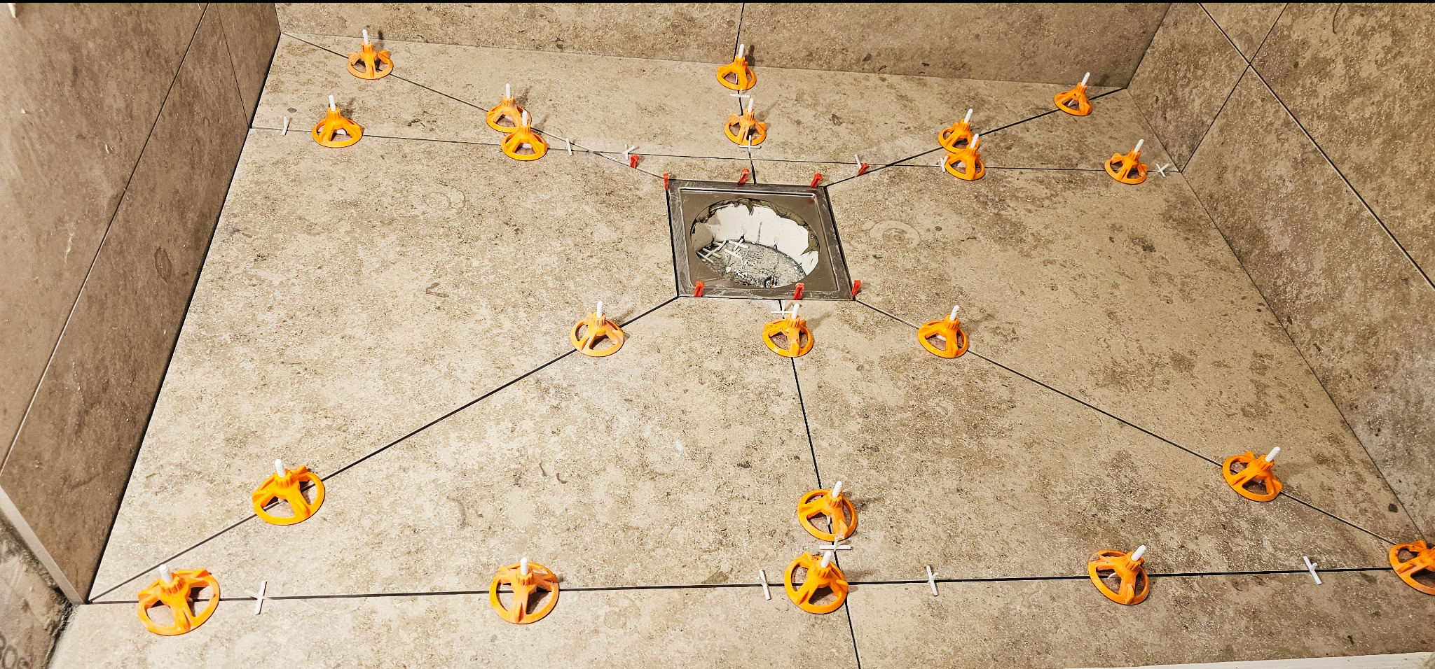 Układanie płytek podłogowych w łazience z użyciem systemu poziomowania. Widoczny odpływ liniowy i pomarańczowe klipsy. Płytki w odcieniach beżu z delikatną strukturą.