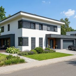 Firma handlowo-usługowa JAKUB RAJ - Nowoczesny dom z szarymi żaluzjami poziomymi na oknach, widok z ulicy w słoneczny dzień. Zadbane otoczenie z zielonym trawnikiem i ozdobnymi roślinami.