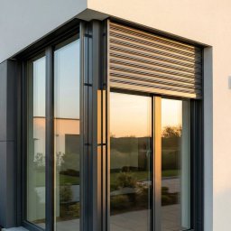 Firma handlowo-usługowa JAKUB RAJ - Nowoczesny dom z oknami zabezpieczonymi roletami zewnętrznymi w kolorze antracytowym, odbijającymi zachód słońca. Eleganckie połączenie bieli i szarości na elewacji.